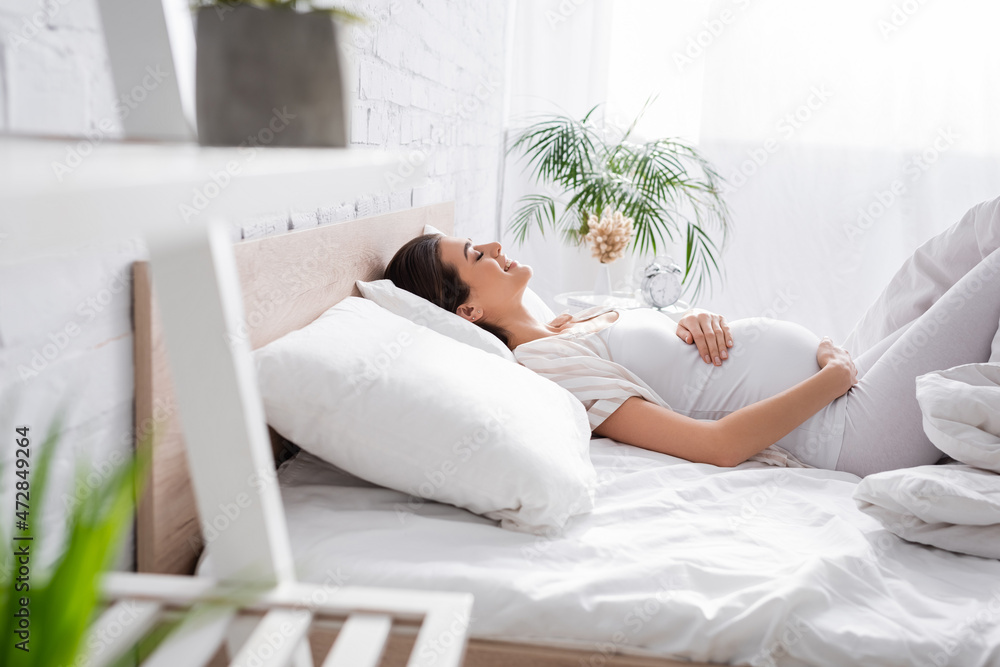 side view of cheerful young pregnant woman touching belly while lying in bedroom. side view of cheerful young pregnant woman touching belly while lying in bedroom.