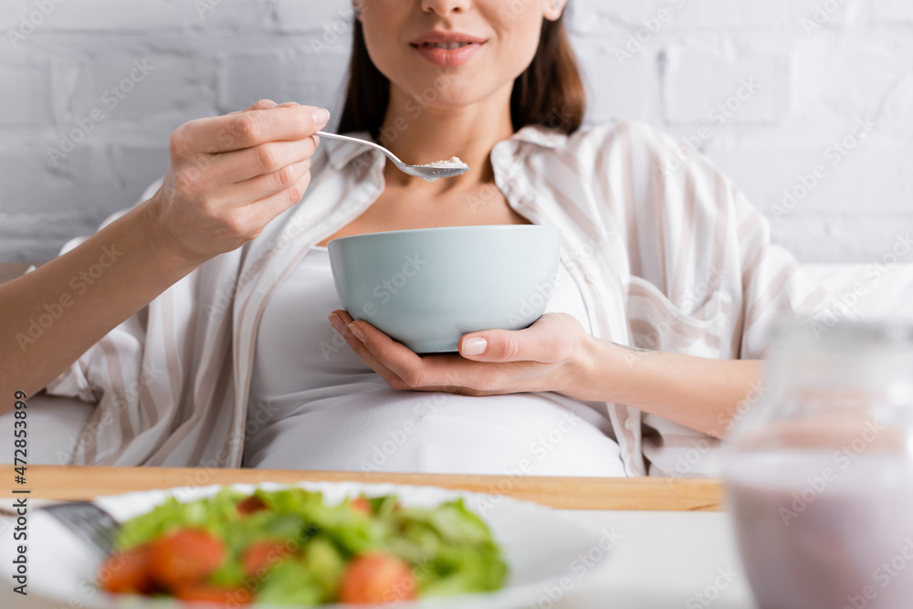 cropped view of happy pregnant woman eating oatmeal near tray with salad. cropped view of happy pregnant woman eating oatmeal near tray with salad.
