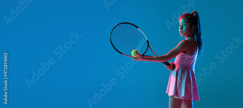 Studio shot of little girl, beginner tennis player training isolated on blue studio background in neon light. Sport, study, childhood concept