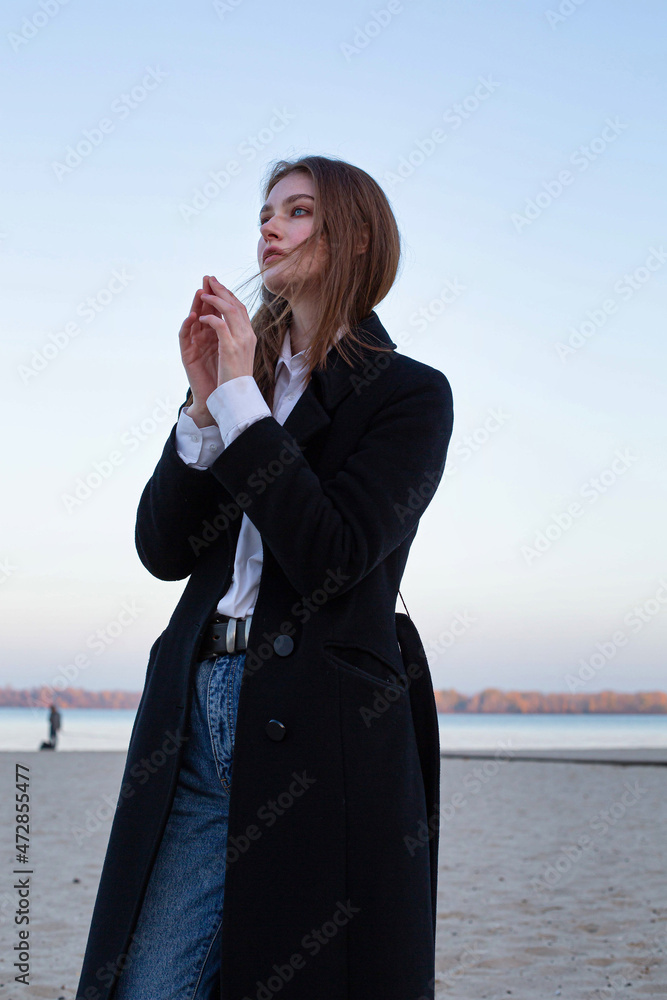 Long straight haired white european woman with folded hands in dark coat in autumn on the beach Long straight haired white european woman with folded hands in dark coat in autumn on the beach