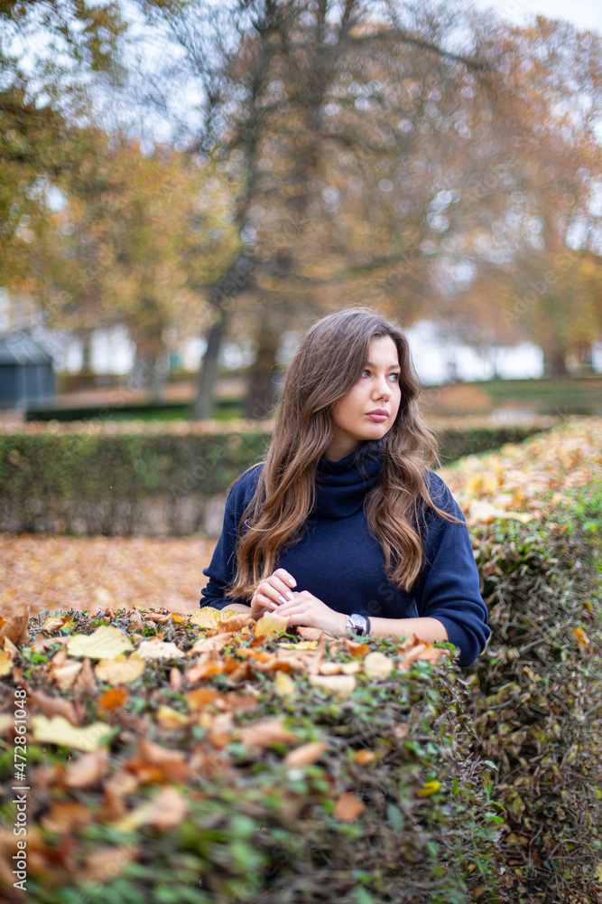 Young european woman in dark blue jumper with long curly hair in autumn park standing near bushes Young european woman in dark blue jumper with long curly hair in autumn park standing near bushes