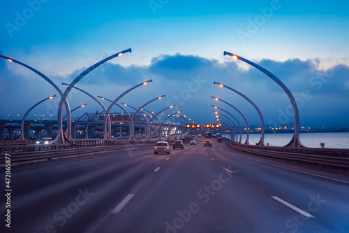 Highways pass over the bridge. Vehicular traffic over the water. Car traffic in the evening. Transport infrastructure of the city. Multi-level highways against the background of the evening city.