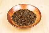 Lot of whole spicy black pepper in ceramic bowl on plain wood
