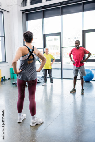 Senior trainer holding hands on hips near blurred multiethnic people in gym.