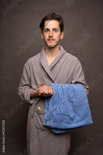 Young handsome tall slim white man with brown hair after shower holding a towel facing the camera in grey bathrobe on grey background