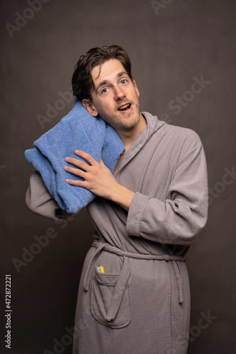 Young handsome tall slim white man with brown hair drying his head with blue towel in grey bathrobe on grey background