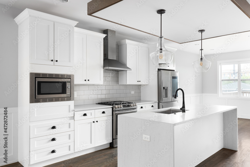 A renovated white kitchen with glass pendant lights hanging above the waterfall granite island, stainless steel appliances, and hardwood floors. A renovated white kitchen with glass pendant lights hanging above the waterfall granite island, stainless steel appliances, and hardwood floors.