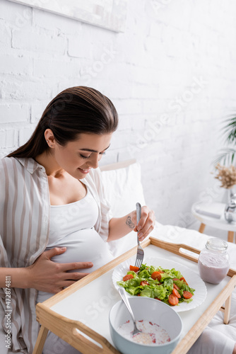 tattooed and happy pregnant woman holding fork near salad on tray.