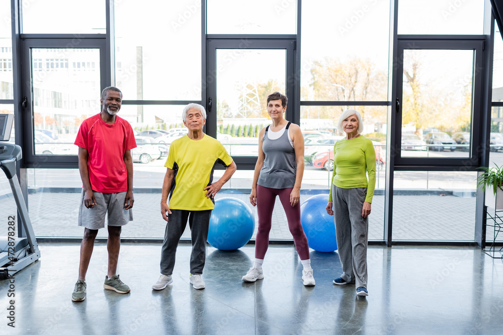 Fototapeta premium Positive multiethnic senior people looking at camera near fitness balls and treadmill in gym.
