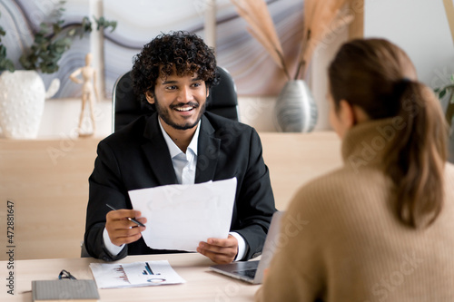 Confident successful Indian male HR manager, entrepreneur, conducts interview girl in a modern office, wearing formal clothes, looking and listening to a job candidate, holding a resume in hands