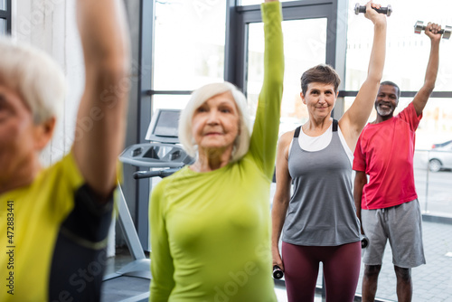 Elderly woman working out with dumbbells near multiethnic friends in gym.