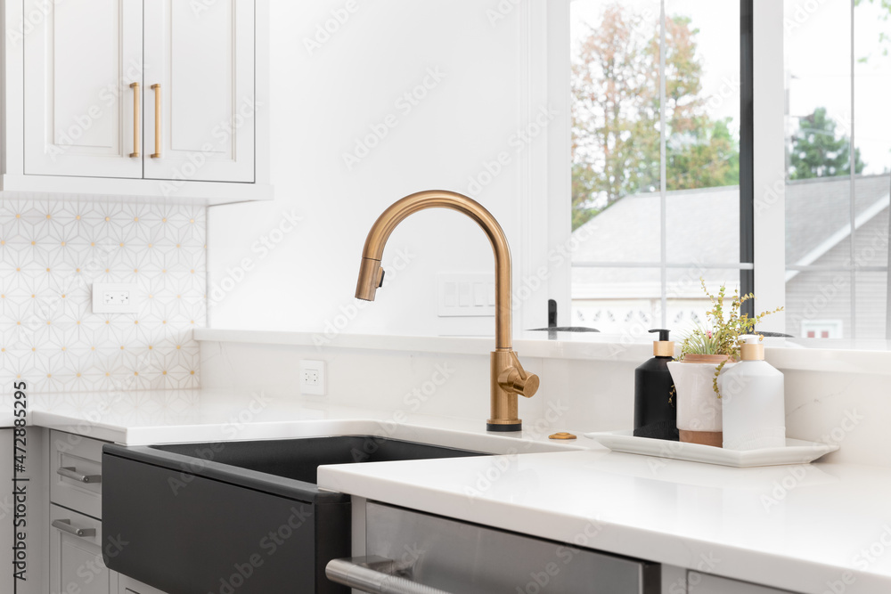A beautiful sink in a remodeled modern farmhouse kitchen with a gold faucet, black apron or farmhouse sink, white granite, and a tiled backsplash. - obrazy, fototapety, plakaty 