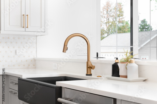 Fotografia A beautiful sink in a remodeled modern farmhouse kitchen with a gold faucet, black apron or farmhouse sink, white granite, and a tiled backsplash