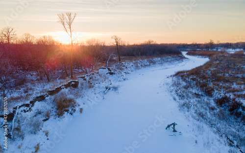 Aerial photo of frozen winding river covered in snow, surrounded by brown vegetation in rural North Dakota. Sun flares created as sun is shining through the trees. Cold, quiet.