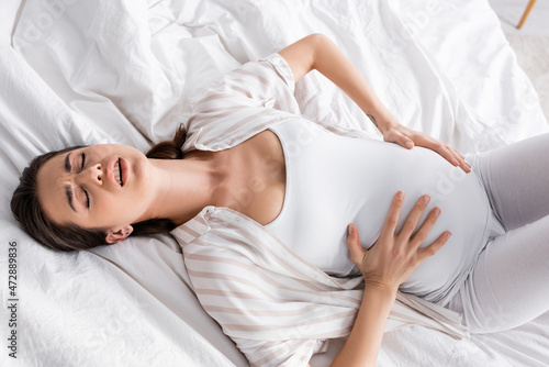 top view of young pregnant woman feeling cramp while lying on bed.