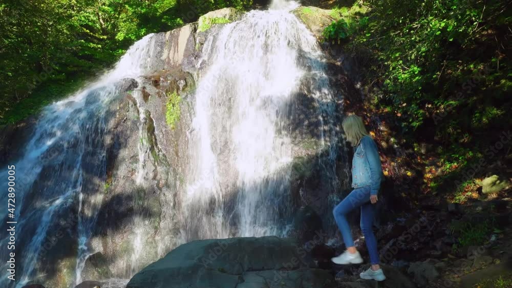 young slender blonde in jeans and a denim jacket, standing on a rock, contemplates a tropical waterfall with its clear clear water in jungle. She raises her hands up and breathes fresh mountain air. young slender blonde in jeans and a denim jacket, standing on a rock, contemplates a tropical waterfall with its clear clear water in jungle. She raises her hands up and breathes fresh mountain air.