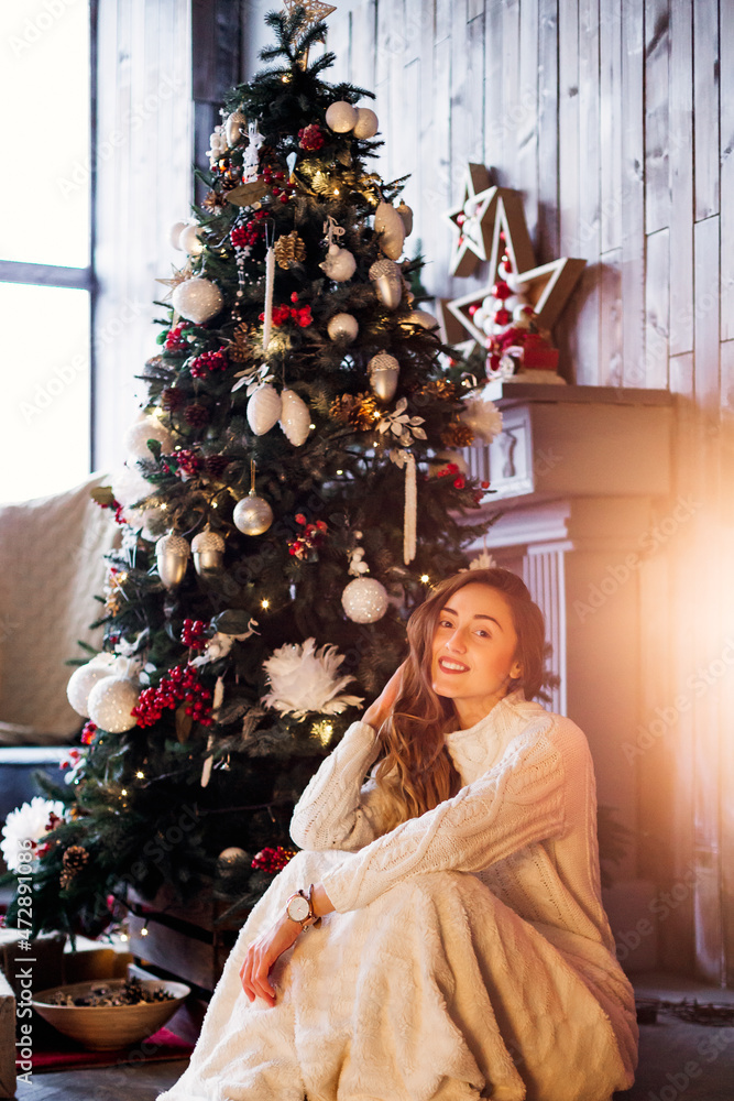 young woman in a spacious living room with New Year's decor sits near a Christmas tree with gifts in a white sweater