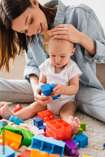 smiling woman touching head of toddler son while playing with building blocks on floor.