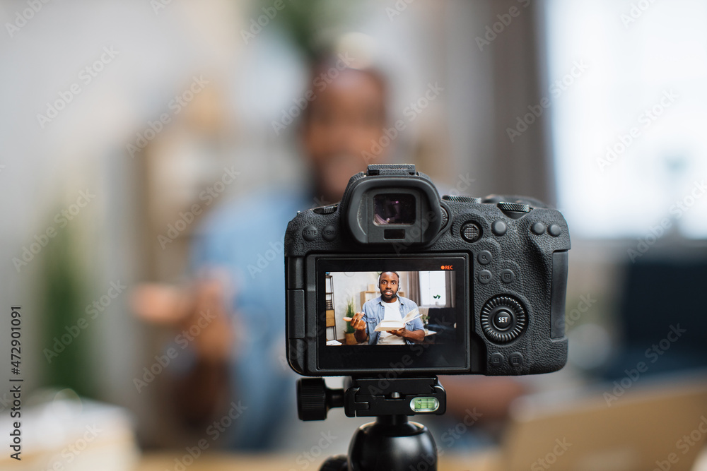 Modern digital camera with african american man on screen. Blur background of smart tutor in headset filming tutorial for students while staying at home.