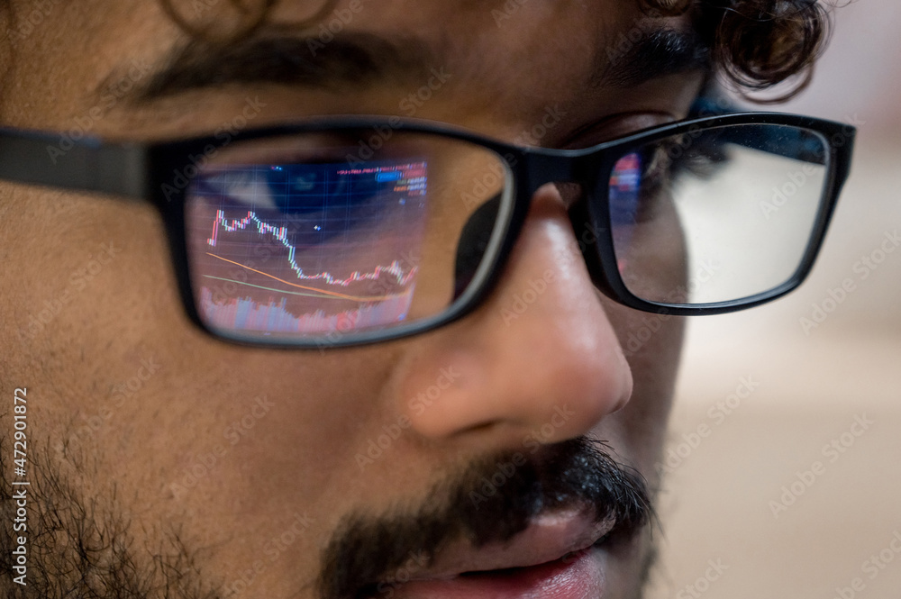 Close-up of Indian man, crypto trader wearing glasses, looking at computer screen with stock exchange market financial data charts reflecting in glasses Close-up of Indian man, crypto trader wearing glasses, looking at computer screen with stock exchange market financial data charts reflecting in glasses