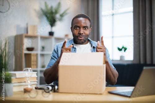 Handsome african american man talking and gesturing on camera while unpacking parcel. First person view of male blogger. Concept of people and social networks.