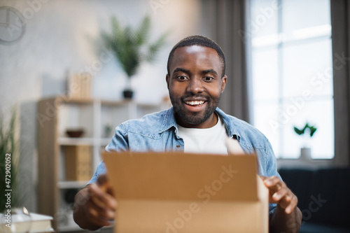 Excited male influencer opening box with ordered goods while staying at home. African american man creating new content for his social networks. photo