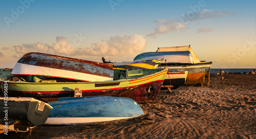 A pile of colorful fisherman boats drying upside down on a sand. Sunset sky panoramic background over coastline of atlantic ocean. Lanzarote, Canary Islands, Spain. Travel destination seascape. © Olga