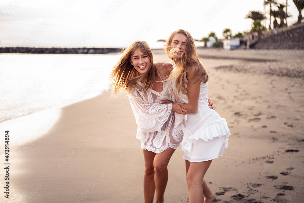 Two young blonde caucasian women walking on the beach, playing and laughing. Female friends having fun on the sea shore. Real people emotions.