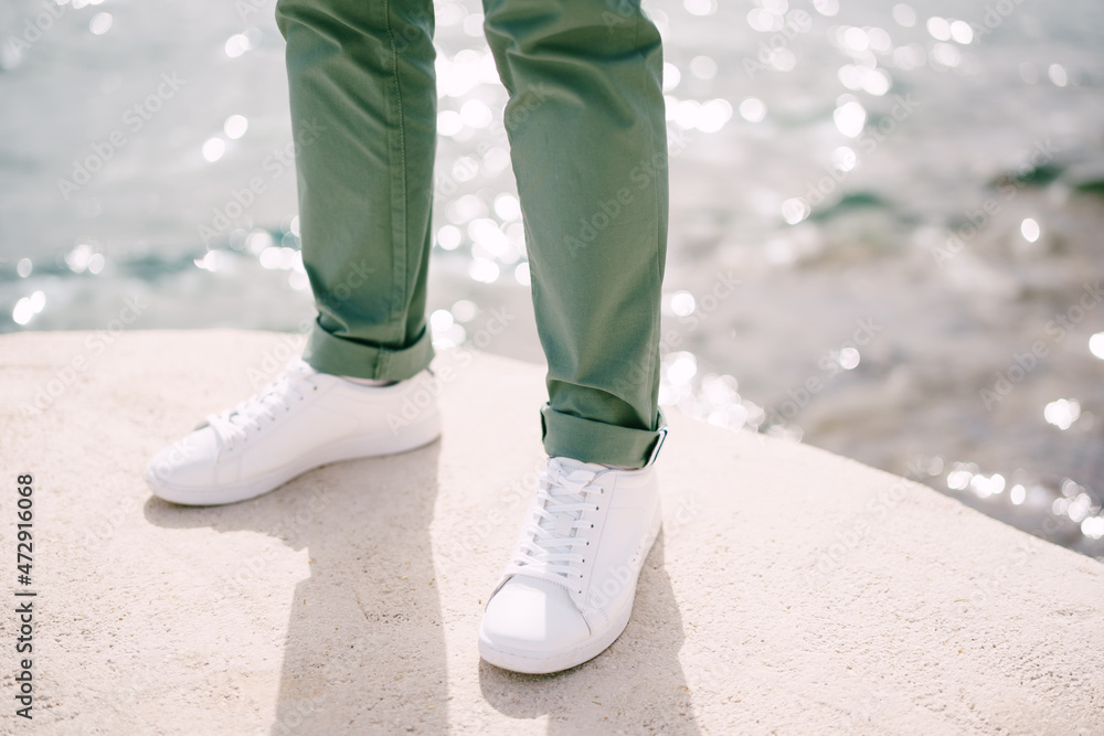 Male feet in white sneakers and green trousers stand on a stone above sparkling water. Close-up