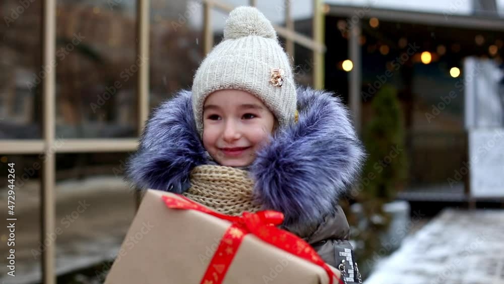 Portrait of a joyful girl with a gift box for Christmas on a city street in winter with snow on a festive market with decorations and lights of garlands. Winter clothes, knitted hat, scarf and fur. Portrait of a joyful girl with a gift box for Christmas on a city street in winter with snow on a festive market with decorations and lights of garlands. Winter clothes, knitted hat, scarf and fur.