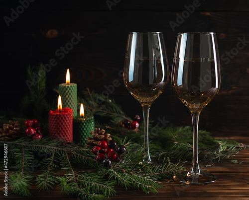 Two glass glasses of champagne on a dark brown wooden table. Red and green candles in the background. Fir branches and glowing garlands on the table. Celebration © Анна Никитина
