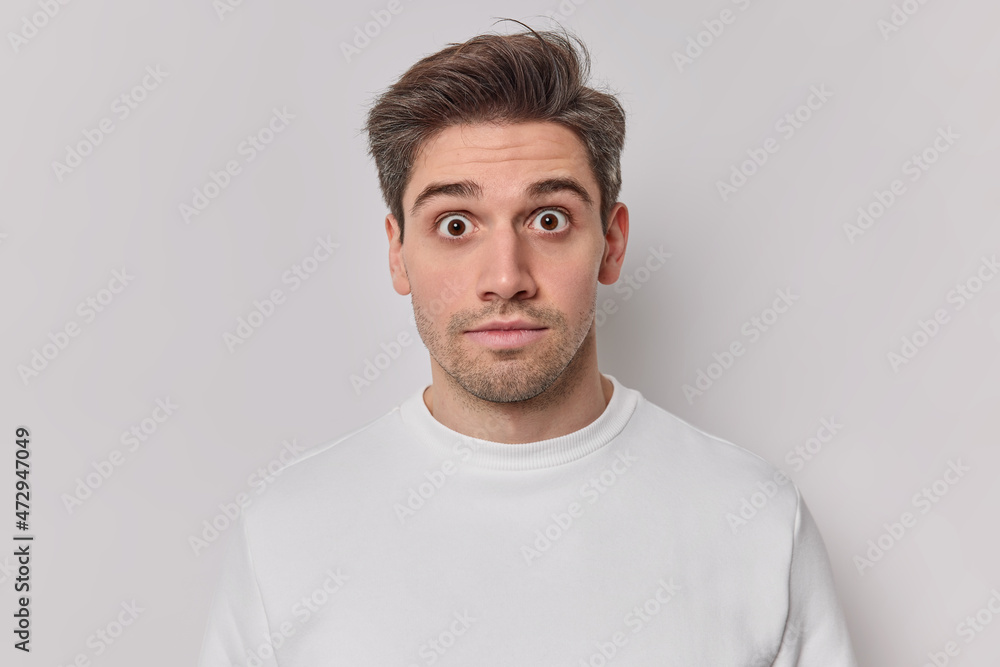 Fototapeta premium Portrait of shocked startled adult man stares bugged eyes at camera hears big news dressed in casual jumper isolated over white background. Concerned young guy feels very surprised poses indoor