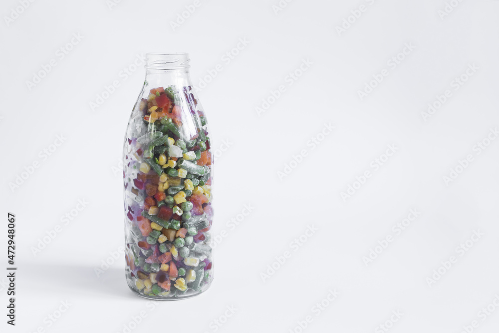 The concept of using quick-frozen products, a transparent bottle with a mixture of frozen vegetables on a light background The concept of using quick-frozen products, a transparent bottle with a mixture of frozen vegetables on a light background