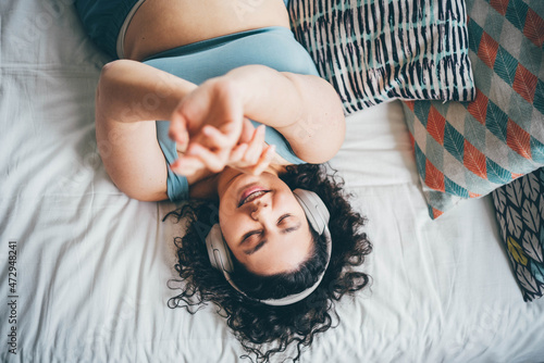 Curly haired plus size young woman in blue top and denim shorts listens to music using wireless headphones and moves hands lying on bed