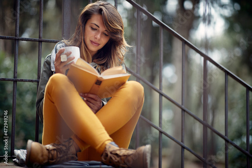 A young girl is focused on a book while sitting on the porch in front of the building and enjoying coffee. City, outside, rest