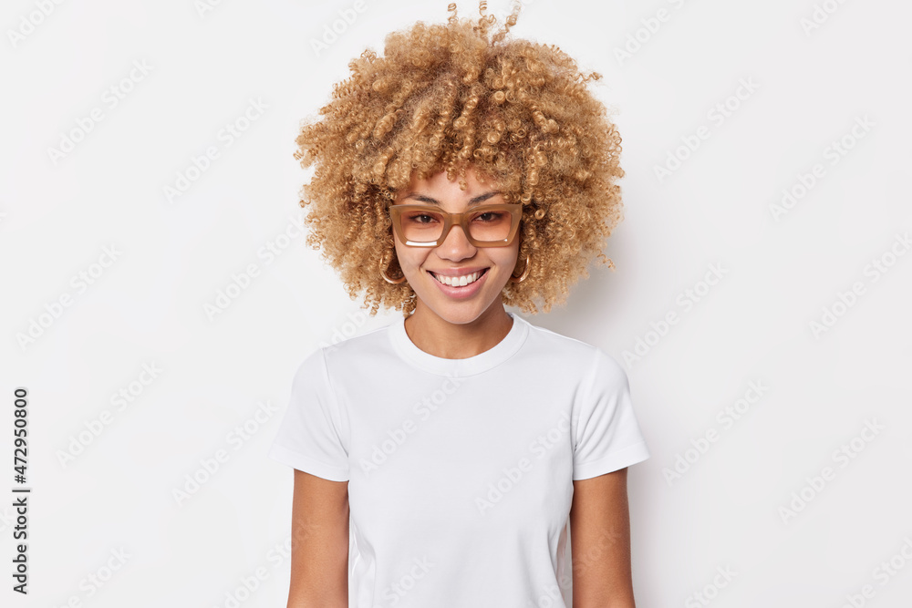 Portrait of pretty young woman with curly hair smiles happily wears spectacles casual basic t shirt has glad expression enjoysfree time poses for making photo isolated over white background. - obrazy, fototapety, plakaty 