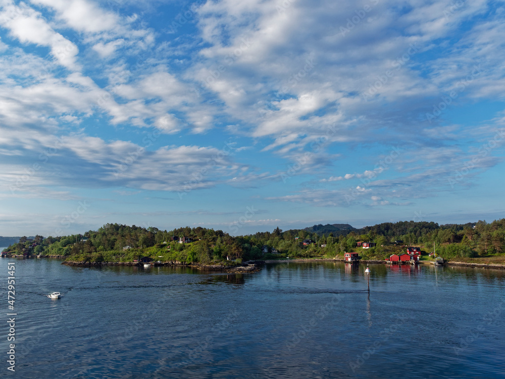 A Small Motorboat cruising past the forested coastline with scattered Houses and Cabins in Bergen Fjord on a bright Summers Morning in June.