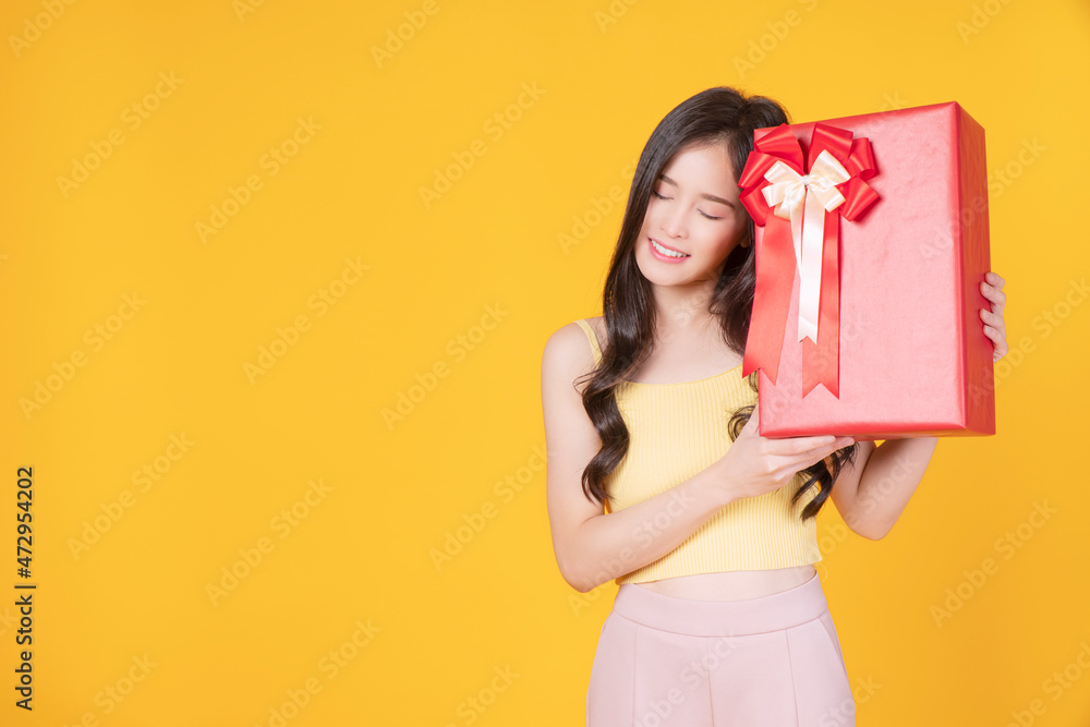Cheerful excited asian woman holding red gift box with ribbon in own hand while standing over isolated blue background. Surprise greeting anniversary birthday or celebration christmas and new year. Cheerful excited asian woman holding red gift box with ribbon in own hand while standing over isolated blue background. Surprise greeting anniversary birthday or celebration christmas and new year.