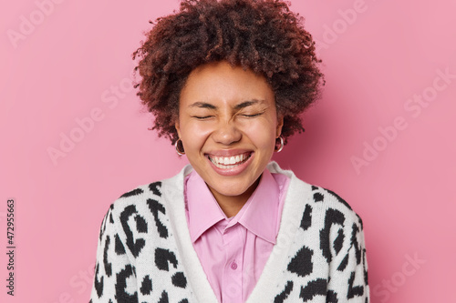 Cuadro en lienzo Positive curly haired woman with curly hair closes eyes and smiles broadly shows white teeth dressed in casual clothes laughs at something funny isolated over pink background