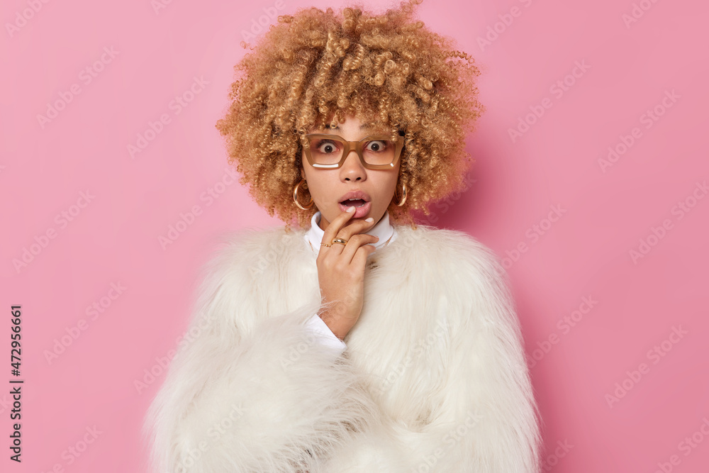Shocked embarrassed young woman with curly hair keeps mouth opened cannot believe in surprising news wears spectacles and white fur coat isolated over pink background. Human reactions concept Shocked embarrassed young woman with curly hair keeps mouth opened cannot believe in surprising news wears spectacles and white fur coat isolated over pink background. Human reactions concept