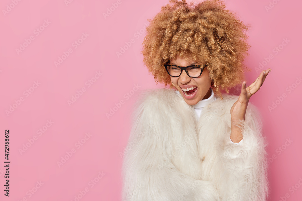 Fashionable woman with curly bushy hair exclaims loudly keeps hand raised reacts on something wears spectacles and white fur coat isolated over pink background with blank copy space for your text Fashionable woman with curly bushy hair exclaims loudly keeps hand raised reacts on something wears spectacles and white fur coat isolated over pink background with blank copy space for your text