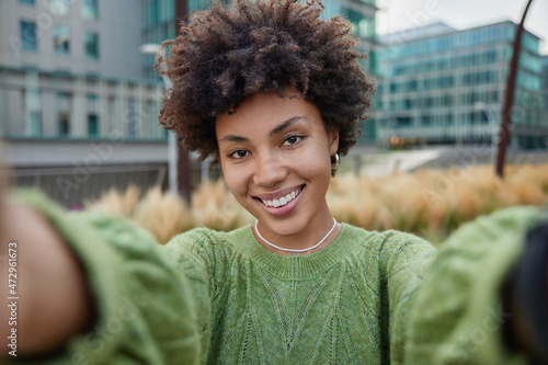 Young pretty woman takes selfie outdoors strolls in street smiles happily wears casual green jumper has happy expression poses against blurred background photographes herself during free time.