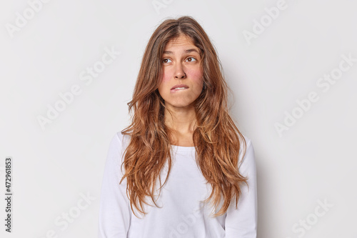 Thoughtful European woman with long hait bites lips considers something has pensive expression dressed in casual jumper isolated over white background makes plan in mind searches for solution