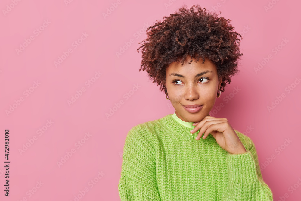 Horizontal shot of thoughtful young woman with curly hair keeps hand under chin concentrated away pensively ponders on something wears knitted sweater isolated over pink background blank space Horizontal shot of thoughtful young woman with curly hair keeps hand under chin concentrated away pensively ponders on something wears knitted sweater isolated over pink background blank space