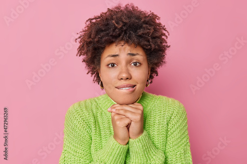 Fotografie Hopeful worried curly haired young woman keeps hands under chin bites lips looks with pleading expression at camera says please or sorry wears warm knitted sweater isolated over pink background