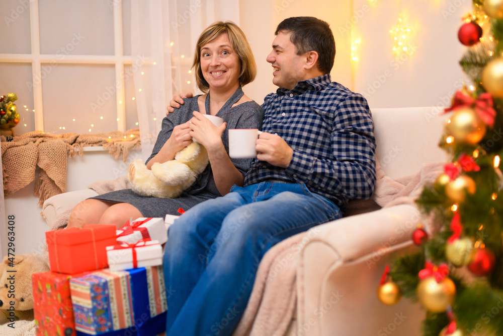 Man and woman sitting on a couch and posing in new year or christmas decoration. They talking and drinking tea or coffee from white cups.. Holiday lights and gifts, Christmas tree decorated with toys. Man and woman sitting on a couch and posing in new year or christmas decoration. They talking and drinking tea or coffee from white cups.. Holiday lights and gifts, Christmas tree decorated with toys.