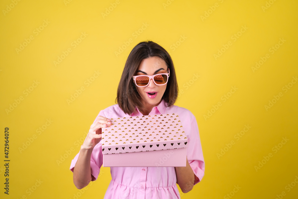 Stylish young woman in a pink dress and glasses on a yellow background with a gift box in her hands celebrates the best holiday, birthday, Valentine's Day Stylish young woman in a pink dress and glasses on a yellow background with a gift box in her hands celebrates the best holiday, birthday, Valentine's Day