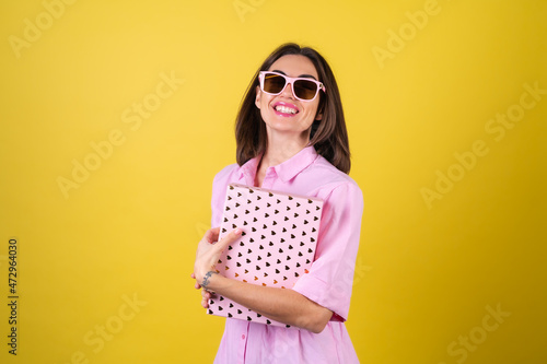 Stylish young woman in a pink dress and glasses on a yellow background with a gift box in her hands celebrates the best holiday, birthday, Valentine's Day