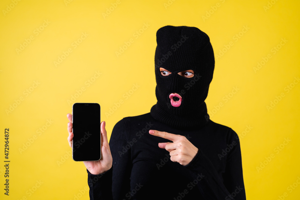 Woman with bright pink lips, balaclava on her head, holding a mobile phone, hacker, internet fraudster, pointing her finger at a blank black screen, excited, shocked Woman with bright pink lips, balaclava on her head, holding a mobile phone, hacker, internet fraudster, pointing her finger at a blank black screen, excited, shocked