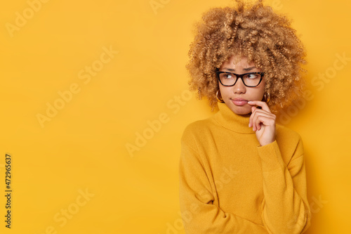 Studio shot of unhappy pensive woman with curly hair keeps hand near corner of lips concentrated into distance wears glasses casual jumper isolated over yellow background copy space for your promotion © Wayhome Studio